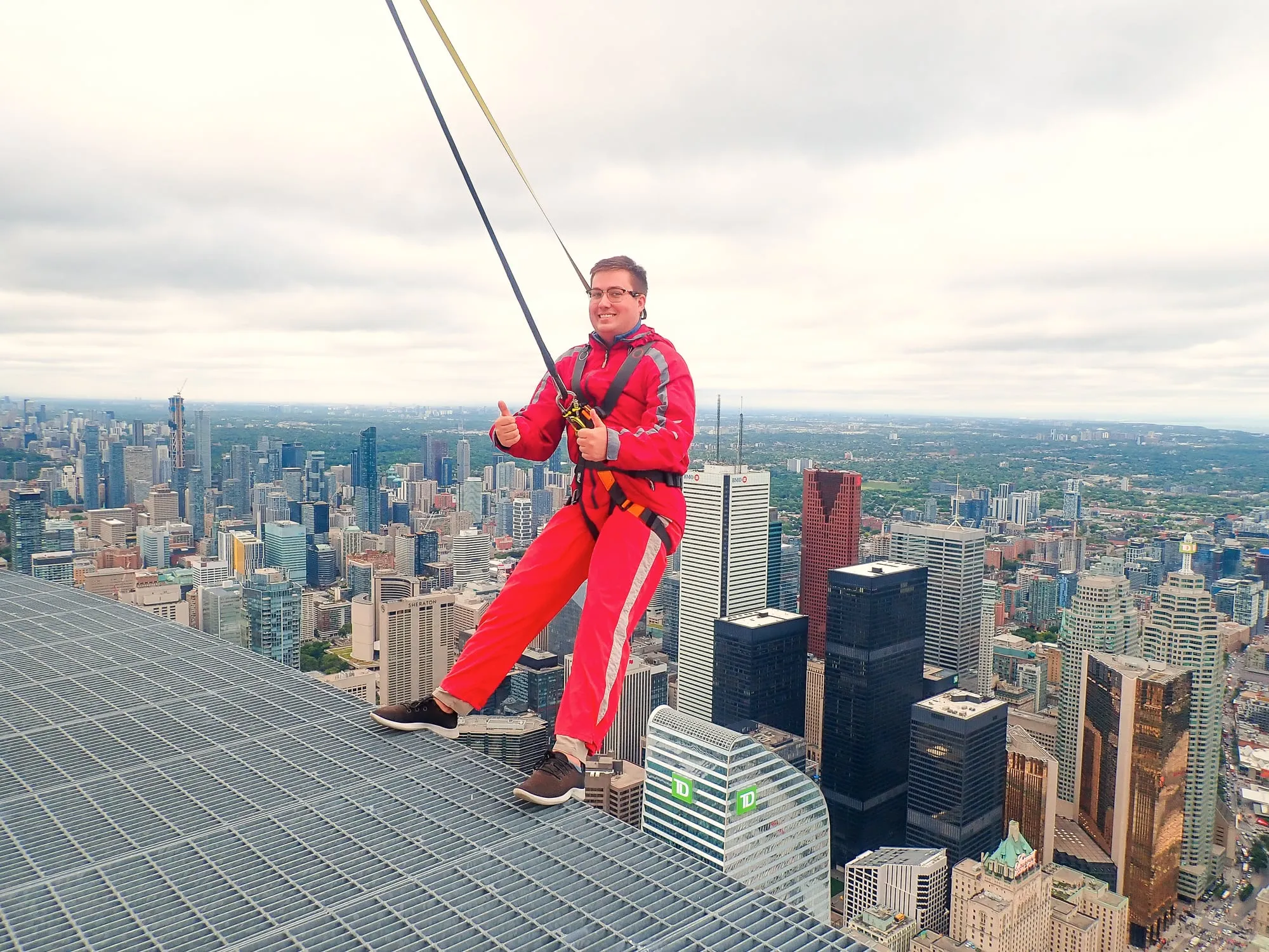 Dominic in a red jumpsuit leaning off the edge of the CN Tower EdgeWalk above downtown Toronto